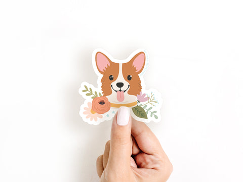 A hand with white nail polish holds the Onderkast Studio Corgi Floral Sticker, a cute, water resistant sticker featuring a corgi with its tongue out among illustrated pink and white flowers on a white background.