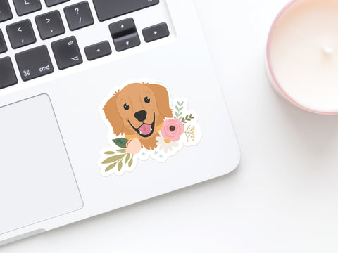 A laptop keyboard with an Onderkast Studio Golden Retriever Floral Sticker—a durable, water-resistant vinyl sticker featuring a smiling cartoon dog and flowers—on the palm rest, beside a lit white candle in a pink holder.