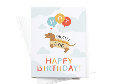 The Hot Diggity Dog Greeting Card by Onderkast Studio shows a cartoon dachshund in a hot dog bun with balloons and a party hat. Premium quality, vibrant HAPPY BIRTHDAY! text—ideal for fun birthday celebrations.