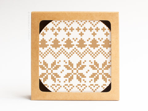 A square brown cardboard box with a window shows an Onderkast Studio Holiday Cross Stitch Coaster, made of durable pulp board and featuring a beige and white pixelated geometric snowflake pattern for lasting handmade quality.