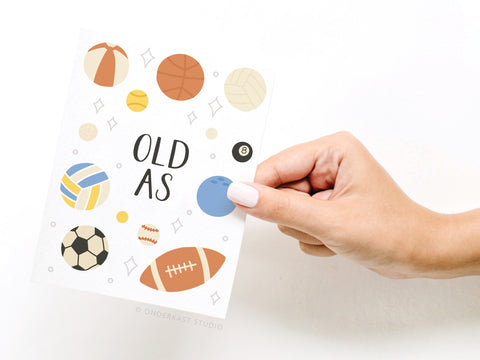 A manicured hand holds the Onderkast Studio Old As Balls Greeting Card, featuring sports balls illustrations and OLD AS text in the center, set against a sleek white background for a premium look.