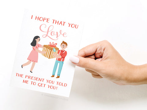 A hand holds the Onderkast Studio I Hope You Love the Present You Told Me To Get You! Greeting Card, featuring a funny woman-giving-gift illustration and bold red text on premium card stock.