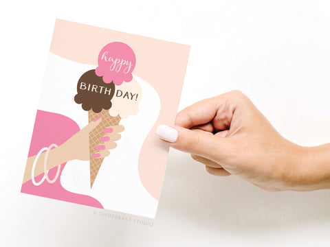 A hand with pink nail polish holds Onderkast Studios Happy Birthday! Ice Cream Cone Greeting Card, featuring scoops labeled happy and BIRTH DAY! against a light background.
