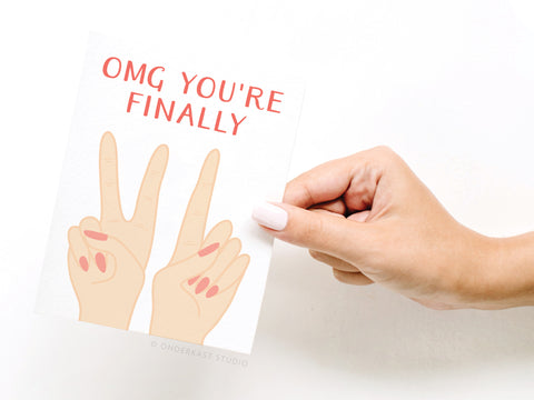 A manicured hand holds the Onderkast Studio OMG Youre Finally [21] Greeting Card, featuring OMG YOURE FINALLY above an illustration of two hands forming the number 21 with their fingers.