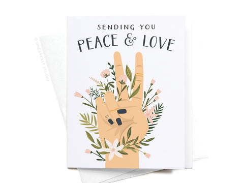 The Sending You Peace & Love Greeting Card by Onderkast Studio features an illustrated peace sign hand with flowers and leaves, plus Sending You Peace & Love text. Light color and a partially visible envelope complete this thoughtful card.